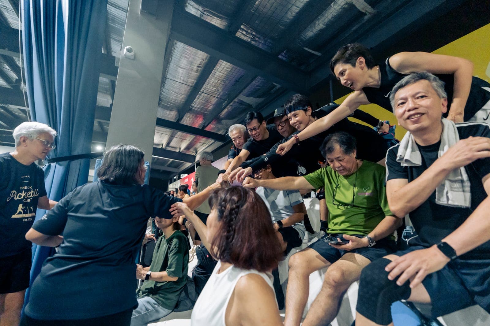 Players and supporters in the stands lean in from every side to join hands and fists in a team huddle on opening night of the Singapore Seniors Pickleball League.